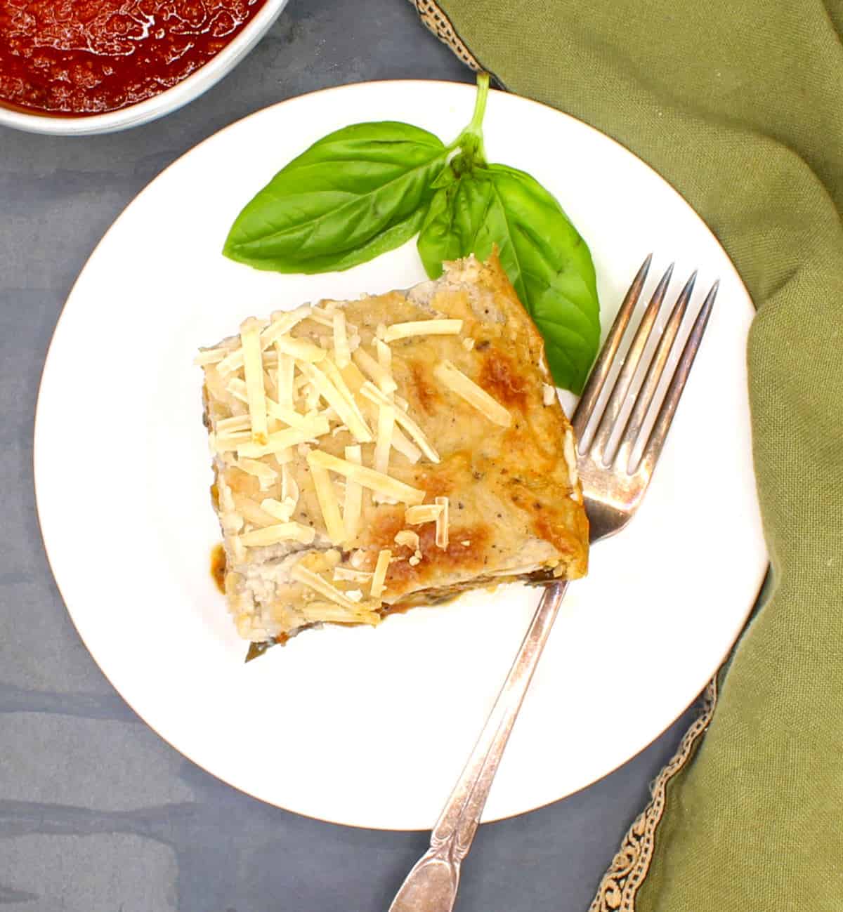 Slice of vegan moussaka with basil leaves in white plate with fork. Tomato sauce in background.