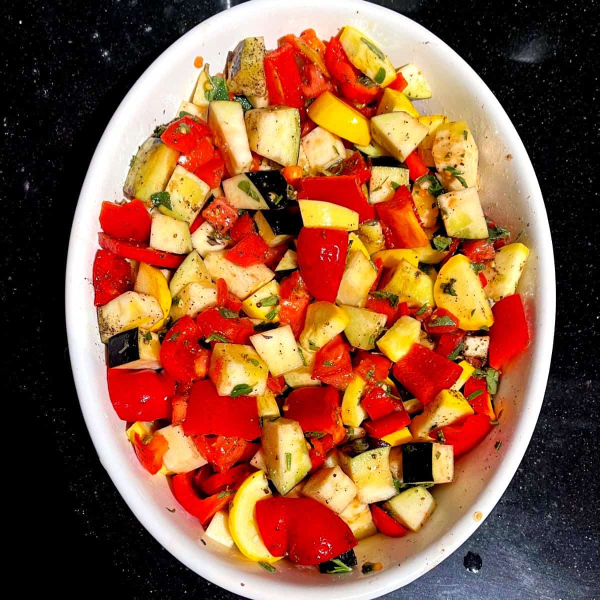 Vegetables for ratatouille mixed with olive oil and herbs in baking dish.