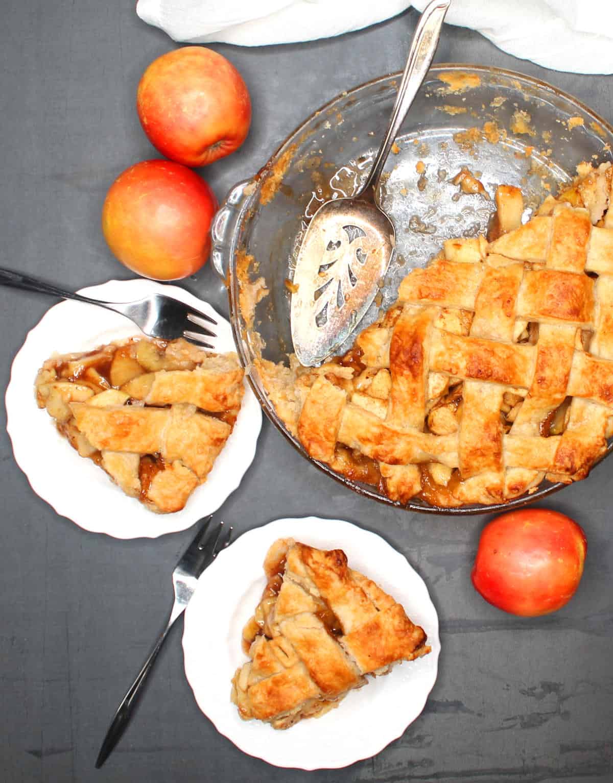 Vegan apple pie with slices of apple pie in white plates and forks and apples scattered around.