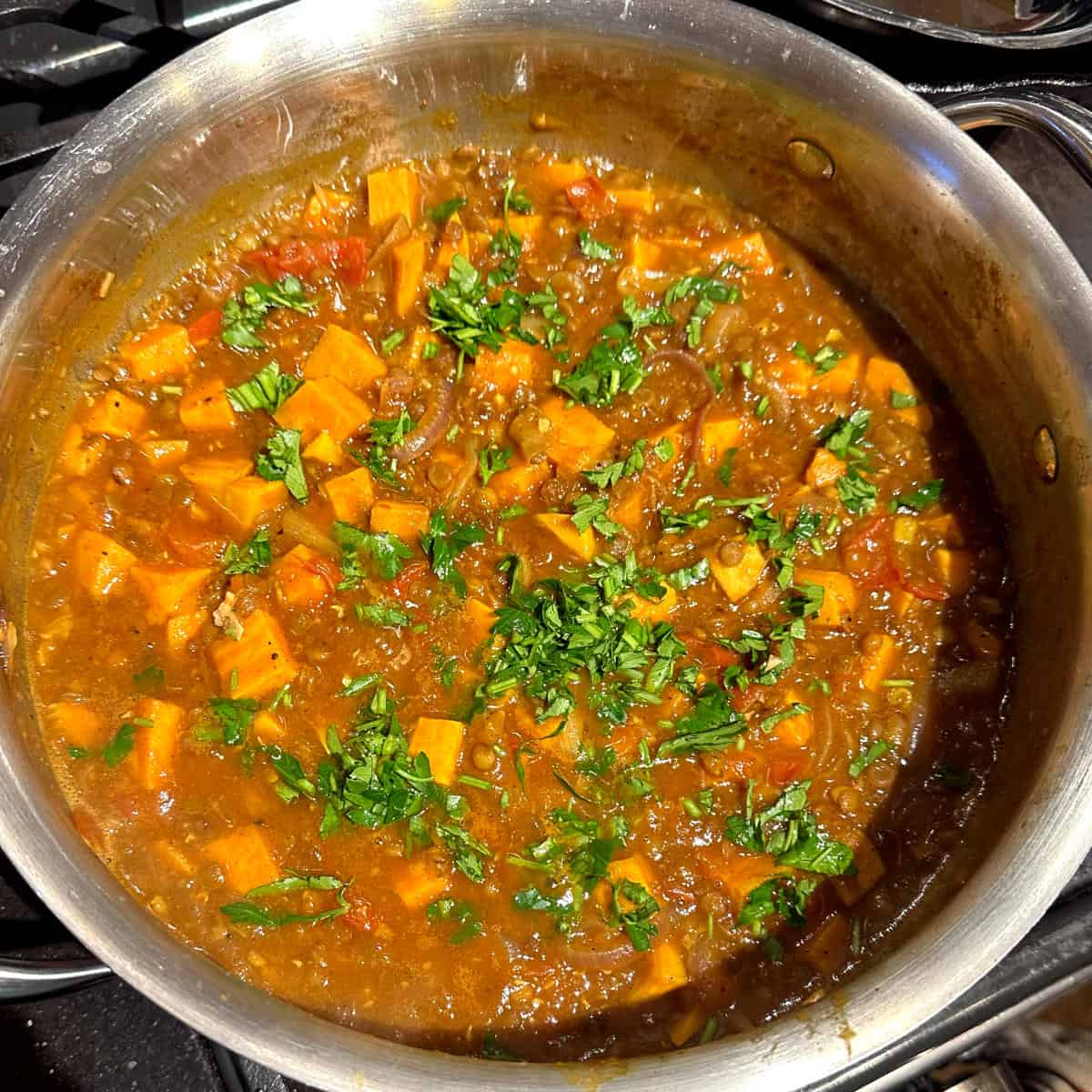Parsley sprinkled over cooked lentil sweet potato tagine in steel pot.