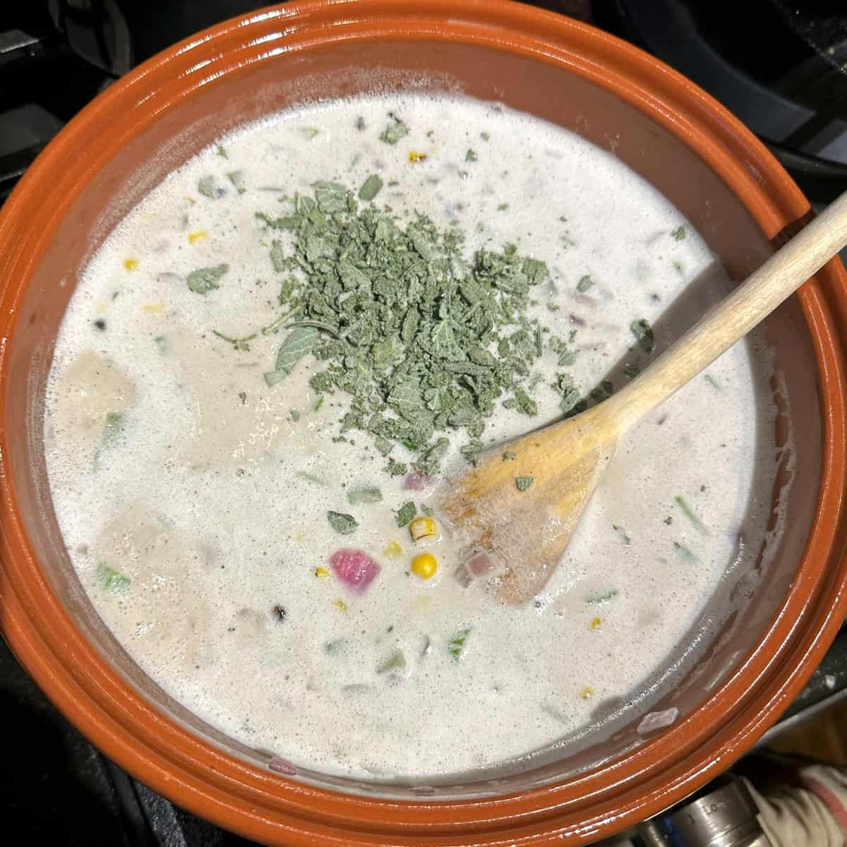 Sage and cumin added to vegan corn chowder in pot.
