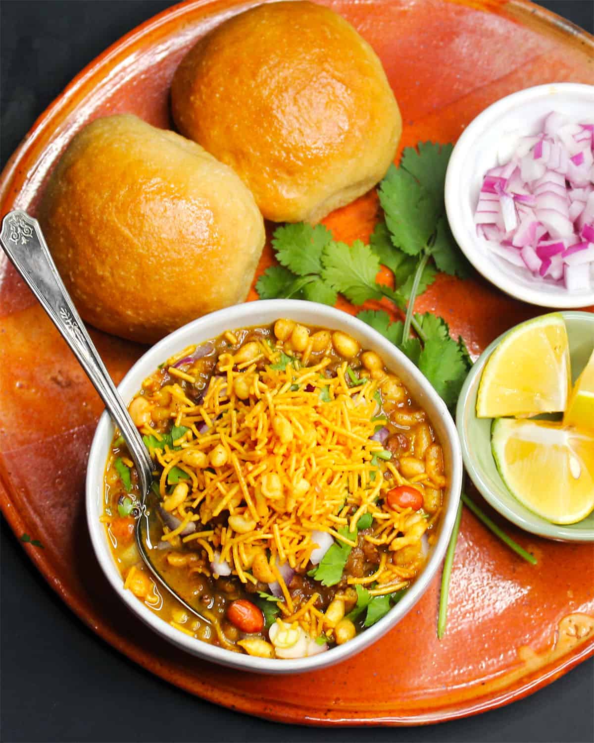 Misal pav in clay platter with onions, lemons and cilantro on the side.