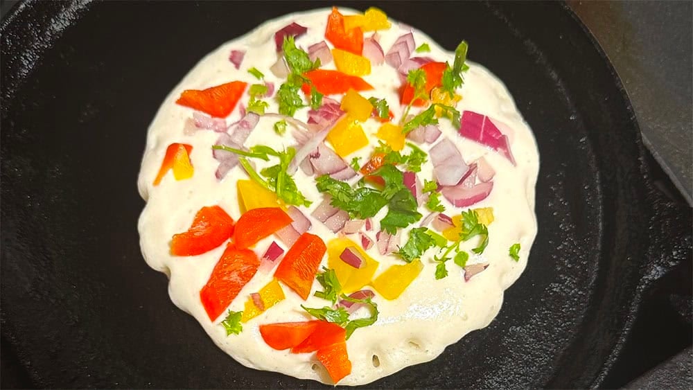 Uttapam batter on skillet with veggies sprinkled on top.