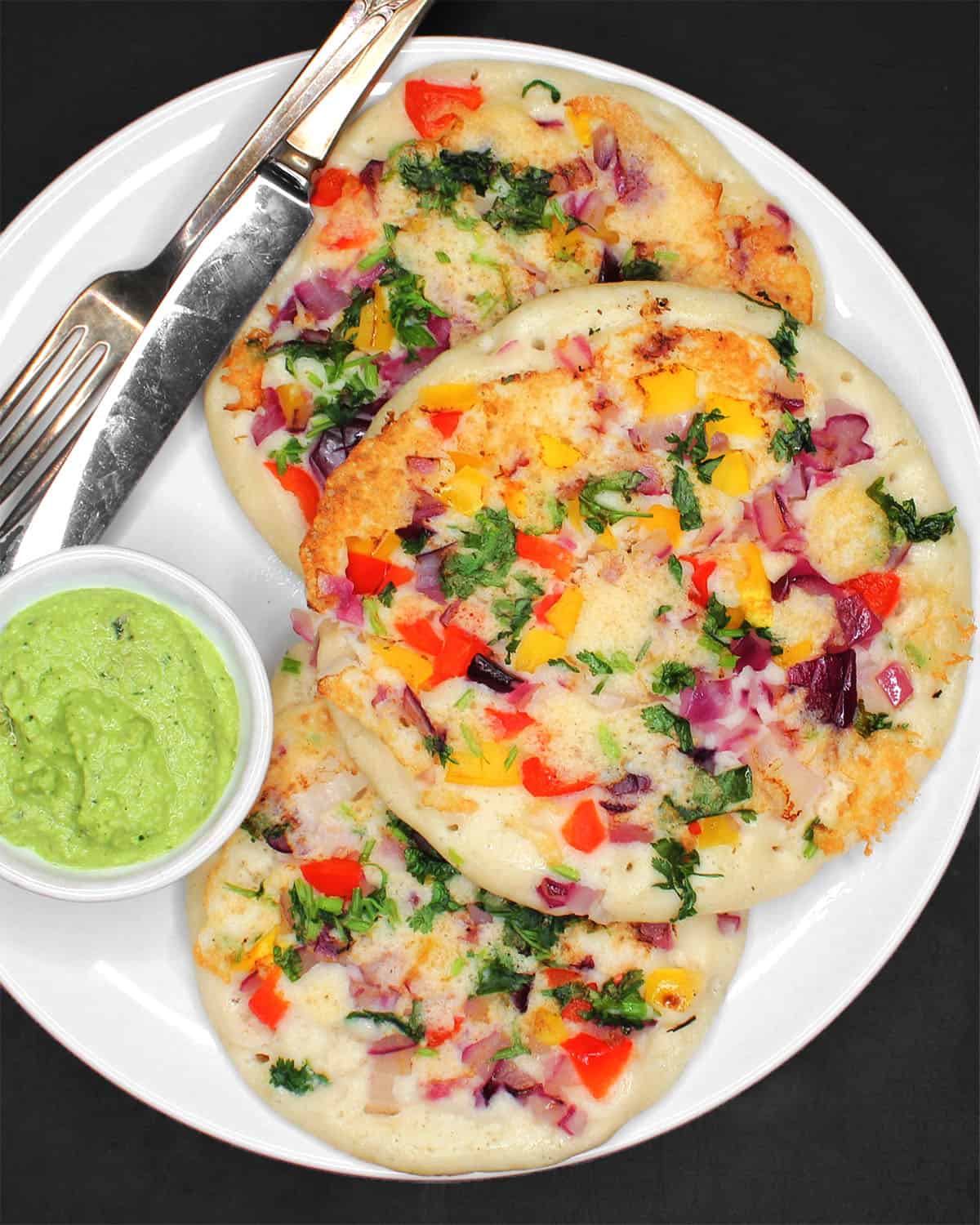 Three fluffy uttapam on plate with coconut chutney, knife and fork.