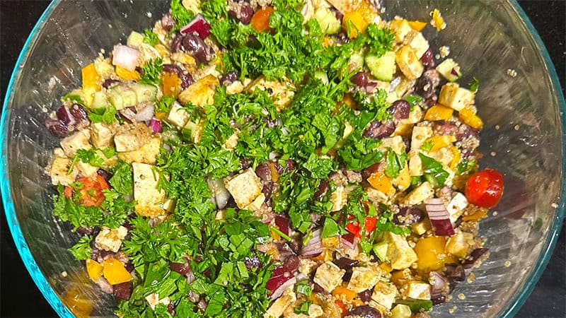 Tofu, quinoa, beans and herbs added to veggies in bowl.