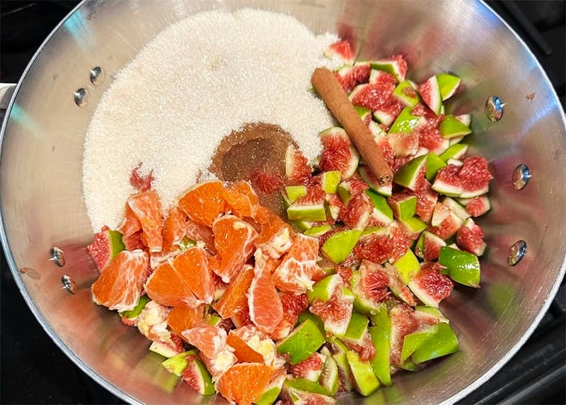 Fig jam ingredients, including figs, sugar, cinnamon, vanilla, lemon juice and orange in steel pan.